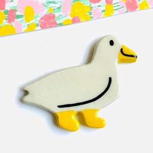 May include: A ceramic duck figurine with a cream-colored body, yellow feet, and a yellow beak. The duck has a black curved line on its side and a black eye. The background includes a colorful patterned paper.