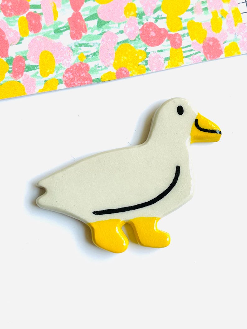 May include: A ceramic duck figurine with a cream-colored body, yellow feet, and a yellow beak. The duck has a black curved line on its side and a black eye. The background includes a colorful patterned paper.
