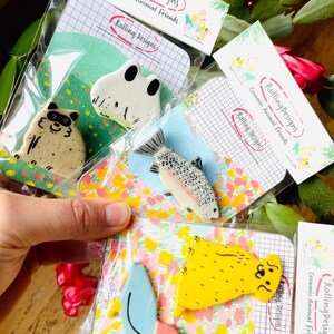 May include: A collection of ceramic animal pins, including a raccoon, frog, and fish, packaged on decorative cards. The pins are colorful and whimsical, with the text "Rolling Designs Ceramic Animal Friends" on the packaging.
