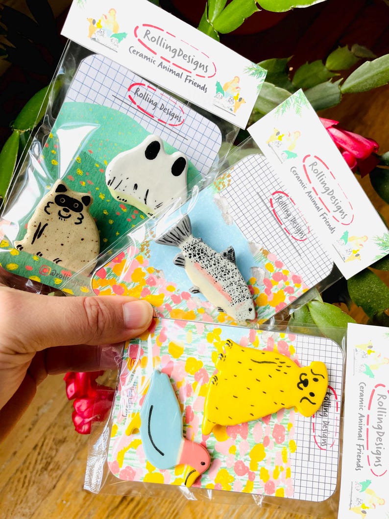 May include: A collection of ceramic animal pins, including a raccoon, frog, and fish, packaged on decorative cards. The pins are colorful and whimsical, with the text "Rolling Designs Ceramic Animal Friends" on the packaging.