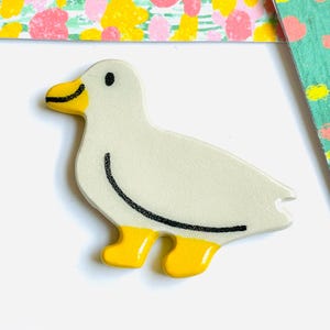 May include: A ceramic duck figurine with a cream-colored body, yellow feet and beak, and black details. The duck has a curved black line on its back and a small black dot for an eye. The background includes floral patterned cards.