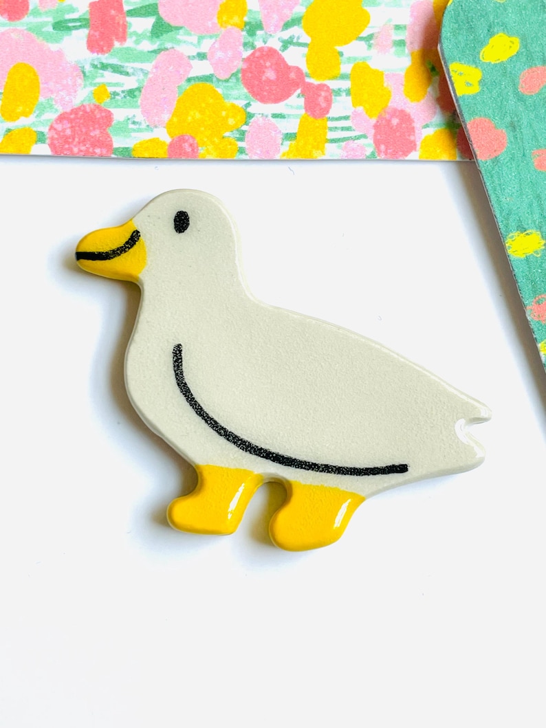 May include: A ceramic duck figurine with a cream-colored body, yellow feet and beak, and black details. The duck has a curved black line on its back and a small black dot for an eye. The background includes floral patterned cards.