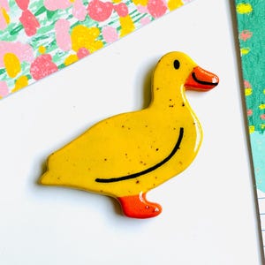 May include: A yellow ceramic duck figurine with a black curved line and small black speckles. The duck has an orange beak and feet. The background includes colorful floral and grid patterns.