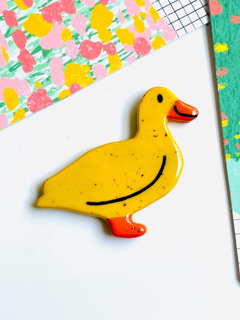 May include: A yellow ceramic duck figurine with a black curved line and small black speckles. The duck has an orange beak and feet. The background includes colorful floral and grid patterns.
