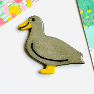 May include: A gray duck-shaped ceramic pin with a yellow beak and feet. The duck has a black outline and a small black eye. The background includes colorful patterned paper.