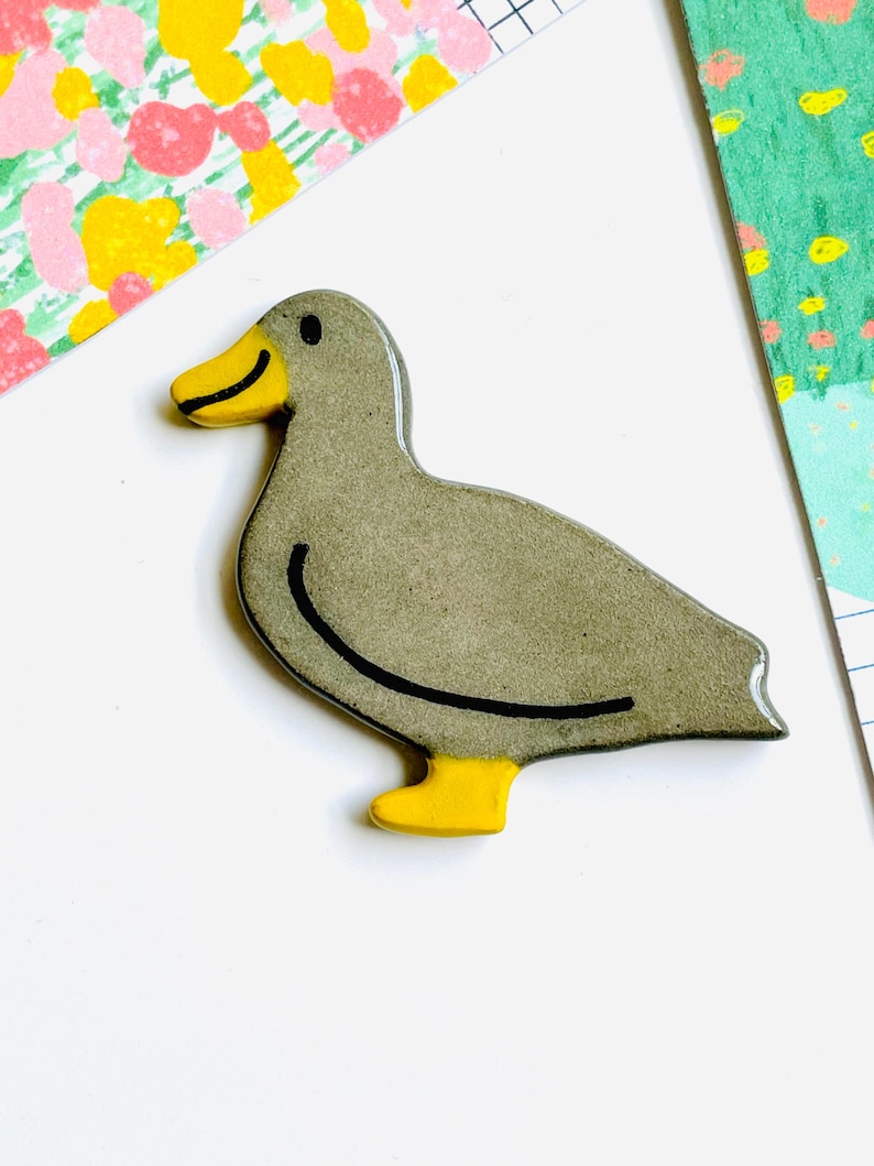 May include: A gray duck-shaped ceramic pin with a yellow beak and feet. The duck has a black outline and a small black eye. The background includes colorful patterned paper.