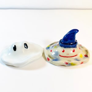 May include: Two ceramic lids with painted faces. One lid is white with black eyes. The other lid is white with blue, red, yellow, and green paint splatters and a blue hat. The lid has a painted smile and red eyes.