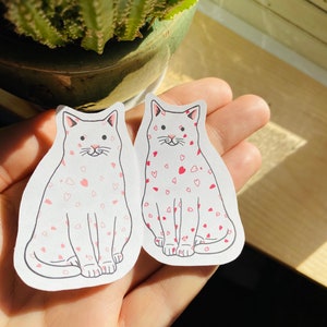 May include: Two white cat stickers with pink hearts on a white background. The cats are sitting with their paws tucked under them and have pink noses and whiskers.