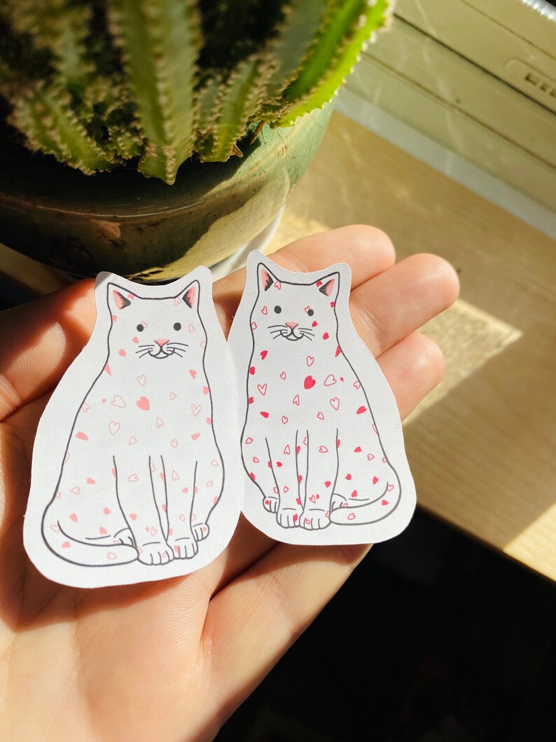 May include: Two white cat stickers with pink hearts on a white background. The cats are sitting with their paws tucked under them and have pink noses and whiskers.