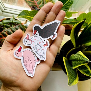 May include: Three duck stickers, one black with red hearts, one white with pink hearts, and one pink with no pattern. The stickers are on a hand.