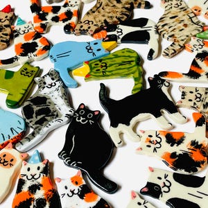 May include: A collection of small, flat, ceramic cat-shaped ornaments featuring various coat patterns including solid black, white, calico, tabby, and spotted designs. Some cats wear small, colorful party hats, and all have hand-painted facial features.