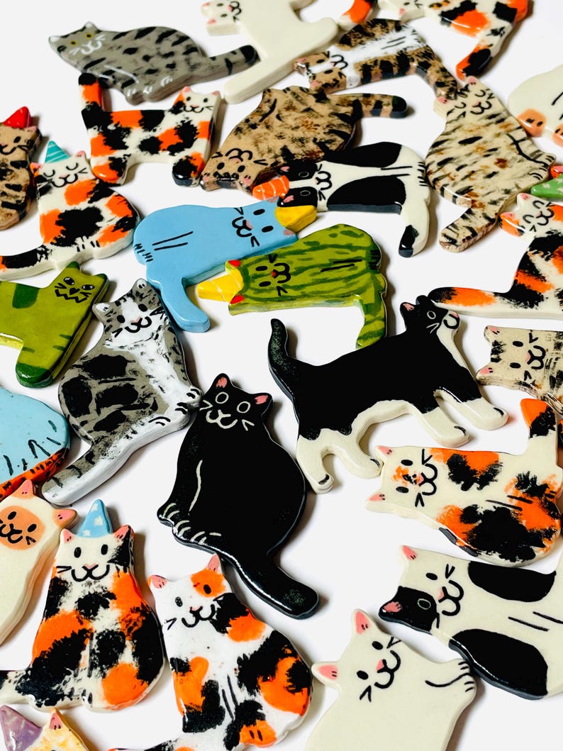 May include: A collection of small, flat, ceramic cat-shaped ornaments featuring various coat patterns including solid black, white, calico, tabby, and spotted designs. Some cats wear small, colorful party hats, and all have hand-painted facial features.