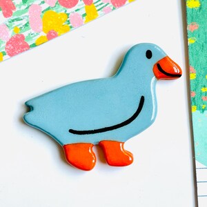 May include: A light blue duck-shaped decorative object with orange feet and beak, and a black curved line for a smile. The duck has a glossy finish and is set against a white background with colorful patterned paper.