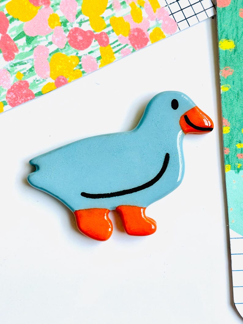 May include: A light blue duck-shaped decorative object with orange feet and beak, and a black curved line for a smile. The duck has a glossy finish and is set against a white background with colorful patterned paper.