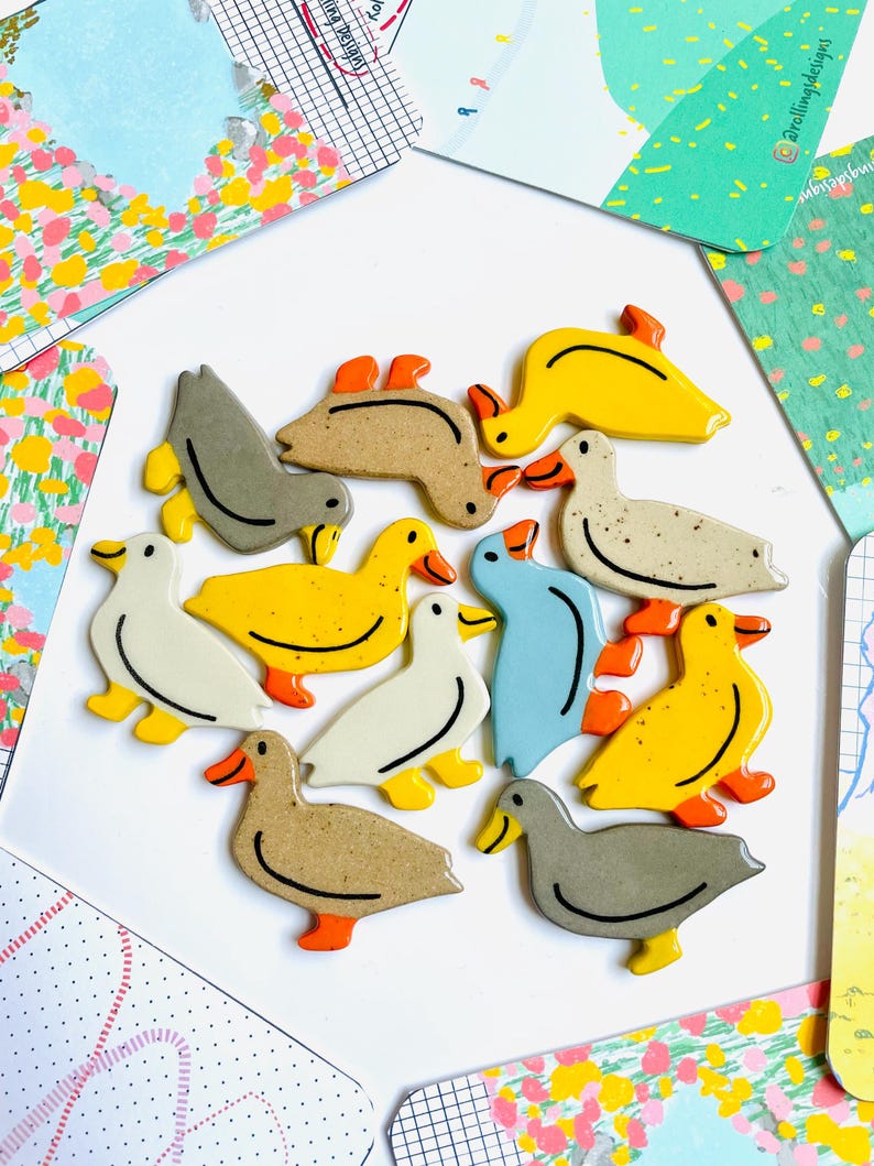 May include: A collection of duck-shaped ceramic magnets in various colors, including yellow, gray, white, and blue, with orange beaks and feet. The magnets are arranged on a white surface, surrounded by colorful cards with floral and abstract designs.