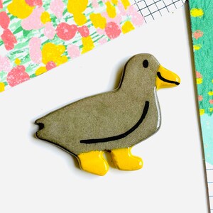 May include: A ceramic duck figurine in shades of gray, with yellow feet and beak. A black curved line accents the duck's body. The duck is set against a white background with colorful patterned paper.