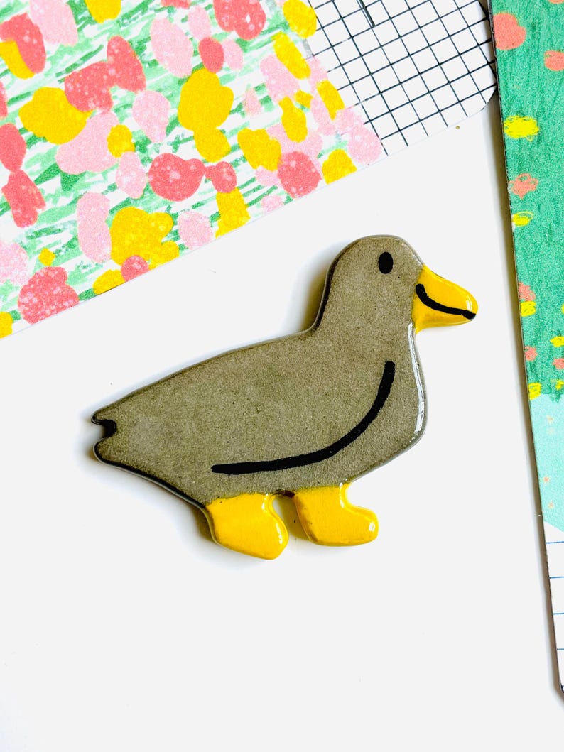 May include: A ceramic duck figurine in shades of gray, with yellow feet and beak. A black curved line accents the duck's body. The duck is set against a white background with colorful patterned paper.