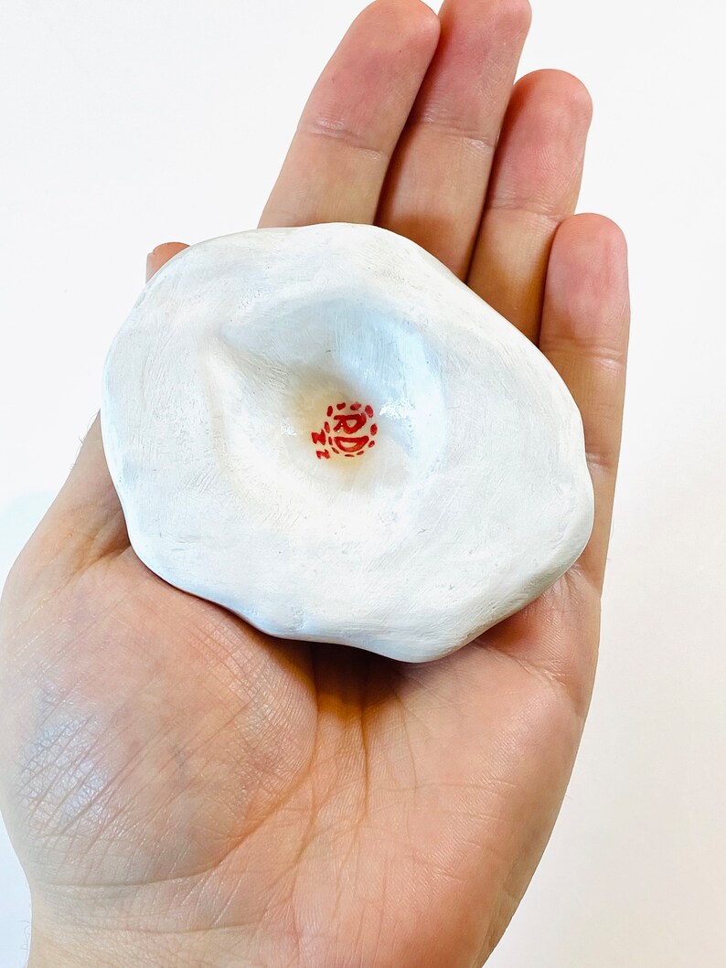May include: White ceramic dish with a deep indentation in the center. The dish has a red stamp with the text "RD" in the center of the indentation.