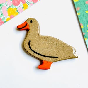 May include: A ceramic duck figurine with a speckled tan body, orange feet and beak, and a black curved line detail. The duck is set against a white background with colorful patterned paper accents.