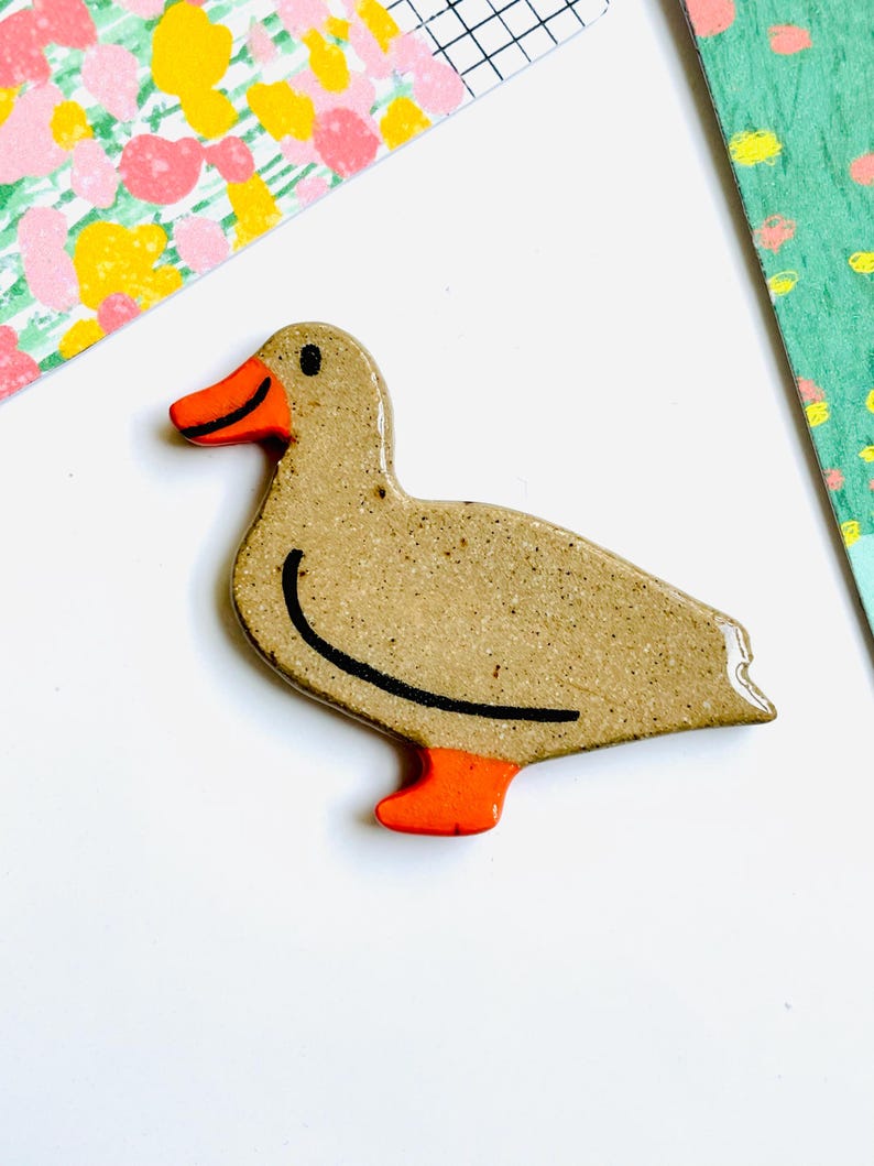 May include: A ceramic duck figurine with a speckled tan body, orange feet and beak, and a black curved line detail. The duck is set against a white background with colorful patterned paper accents.