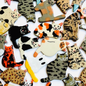 Può includere: Una collezione di statuine di gatto in ceramica in vari colori e motivi, tra cui tartarugato, tabby e bianco e nero. Ogni gatto ha un design unico, con dettagli come accenti rosa o blu sulle orecchie e sulle zampe.