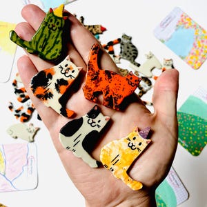 May include: A collection of small, flat, ceramic cat-shaped pins featuring various patterns and colors, including a green striped cat, a calico cat, a bright orange mottled cat, a black and white cat, and a yellow cat with a purple party hat.