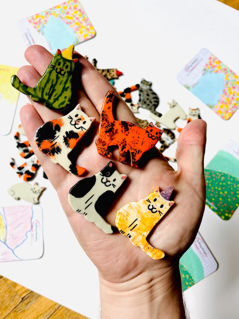 May include: A collection of small, flat, ceramic cat-shaped pins featuring various patterns and colors, including a green striped cat, a calico cat, a bright orange mottled cat, a black and white cat, and a yellow cat with a purple party hat.
