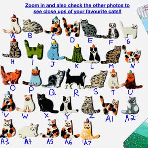 May include: A collection of thirty-four small, flat, ceramic cat-shaped pins or ornaments, each labeled with a letter or alphanumeric code from A to A7. The cats feature various colors, patterns, and some wear small, colorful party hats.