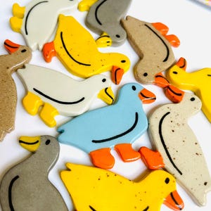 May include: A collection of ceramic duck figurines in various colors, including yellow, blue, gray, and beige. Each duck has orange feet and beaks, with black detailing. The ducks are arranged on a white surface.