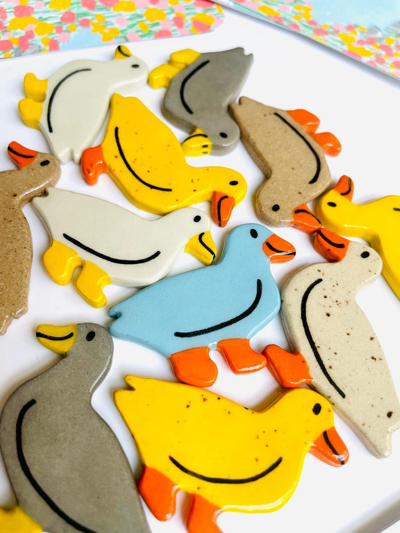 May include: A collection of ceramic duck figurines in various colors, including yellow, blue, gray, and beige. Each duck has orange feet and beaks, with black detailing. The ducks are arranged on a white surface.