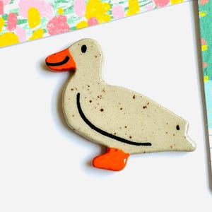 May include: A ceramic duck figurine with a speckled, off-white body, orange beak and feet, and a black curved line for the back. The duck is on a white surface with colorful patterned paper in the background.