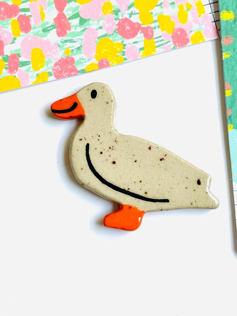 May include: A ceramic duck figurine with a speckled, off-white body, orange beak and feet, and a black curved line for the back. The duck is on a white surface with colorful patterned paper in the background.