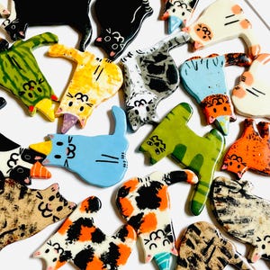 May include: A collection of flat, ceramic cat-shaped ornaments in various colors and patterns, including calico, tabby, solid black, and green. Several cats feature small, colorful party hats on their heads, and all display painted facial features.