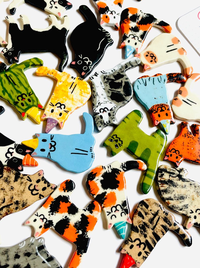 May include: A collection of flat, ceramic cat-shaped ornaments in various colors and patterns, including calico, tabby, solid black, and green. Several cats feature small, colorful party hats on their heads, and all display painted facial features.