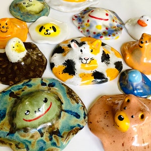 May include: A collection of 12 ceramic figurines, each depicting a different animal with a unique personality. The figurines are painted in bright colors and have a whimsical, cartoonish style. They are all shaped like a fried egg, with the yolk being the animal's head.