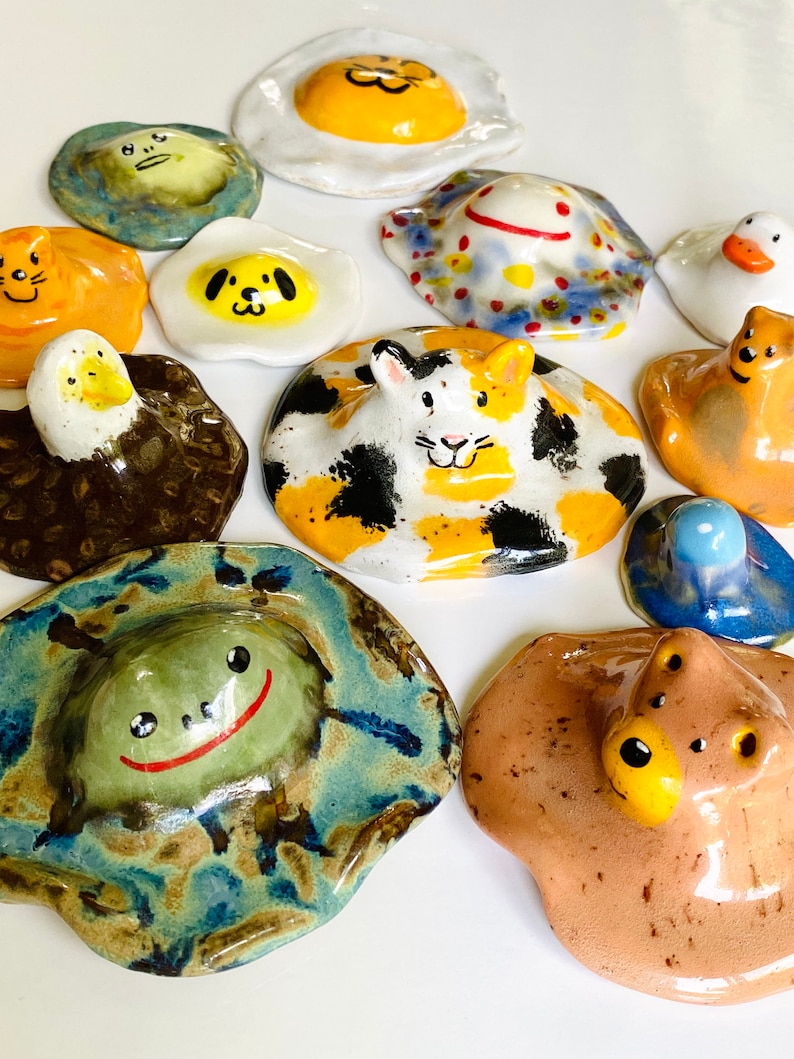 May include: A collection of 12 ceramic figurines, each depicting a different animal with a unique personality. The figurines are painted in bright colors and have a whimsical, cartoonish style. They are all shaped like a fried egg, with the yolk being the animal's head.
