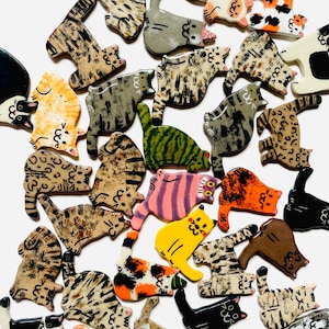 May include: A collection of small, flat, cat-shaped ceramic ornaments featuring various coat patterns including tabby, calico, solid black, white, orange, and spotted designs. Each piece displays a hand-painted feline silhouette with simple facial features.