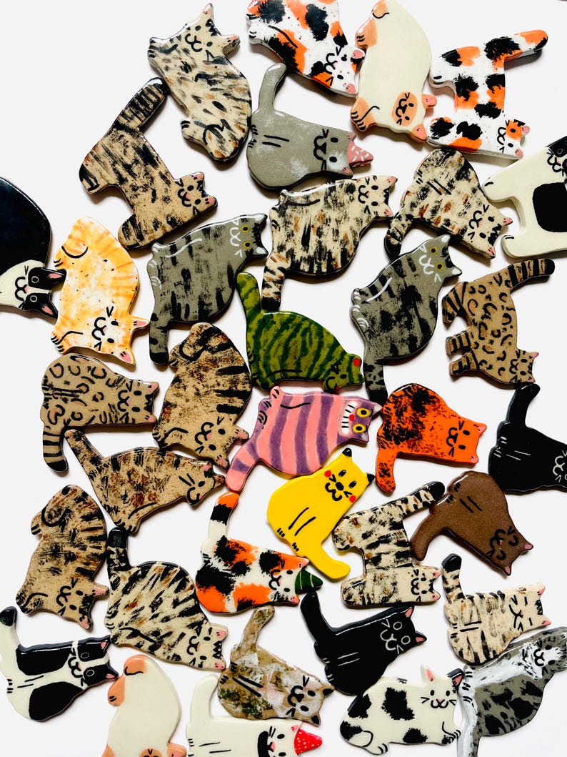 May include: A collection of small, flat, cat-shaped ceramic ornaments featuring various coat patterns including tabby, calico, solid black, white, orange, and spotted designs. Each piece displays a hand-painted feline silhouette with simple facial features.