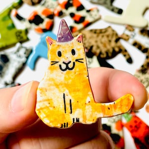 May include: A small, flat, ceramic cat figurine with a glossy orange and yellow finish, featuring a purple party hat, black hand-painted facial details, and simple vertical lines representing legs, held between fingers against a blurred background.