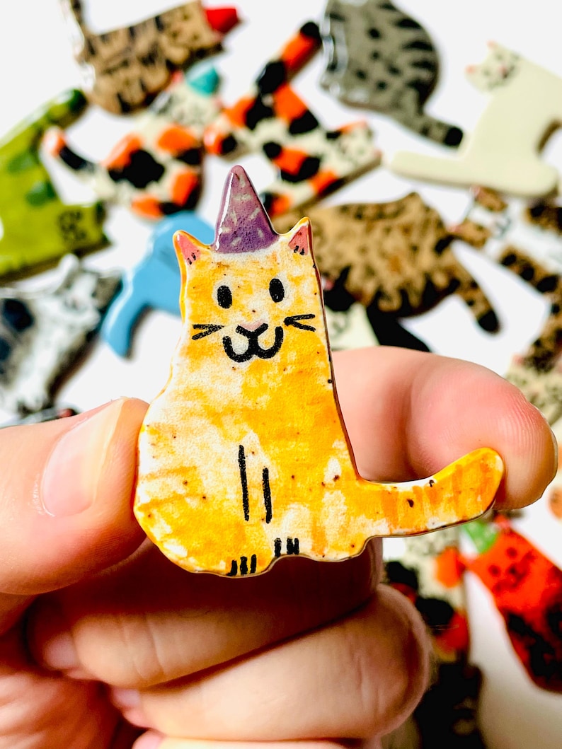 May include: A small, flat, ceramic cat figurine with a glossy orange and yellow finish, featuring a purple party hat, black hand-painted facial details, and simple vertical lines representing legs, held between fingers against a blurred background.