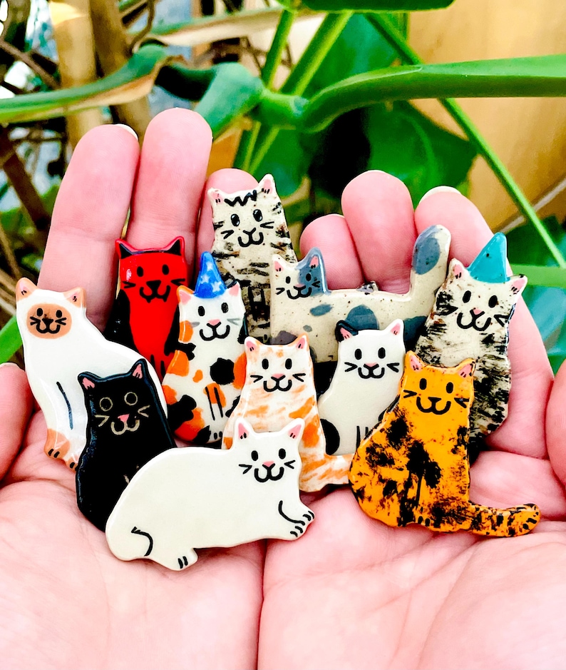 May include: A collection of small, flat, ceramic cat-shaped ornaments in various colors and patterns, including solid black, white, orange, and calico designs. Some cats feature painted details like party hats, stripes, and spots with simple faces.