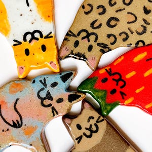 May include: Five ceramic cat-shaped ornaments with glossy finishes. One is white and orange, one is tan with black spots, one is blue and orange, one is red with yellow streaks, and one is brown with darker brown patches, all featuring black line art faces.