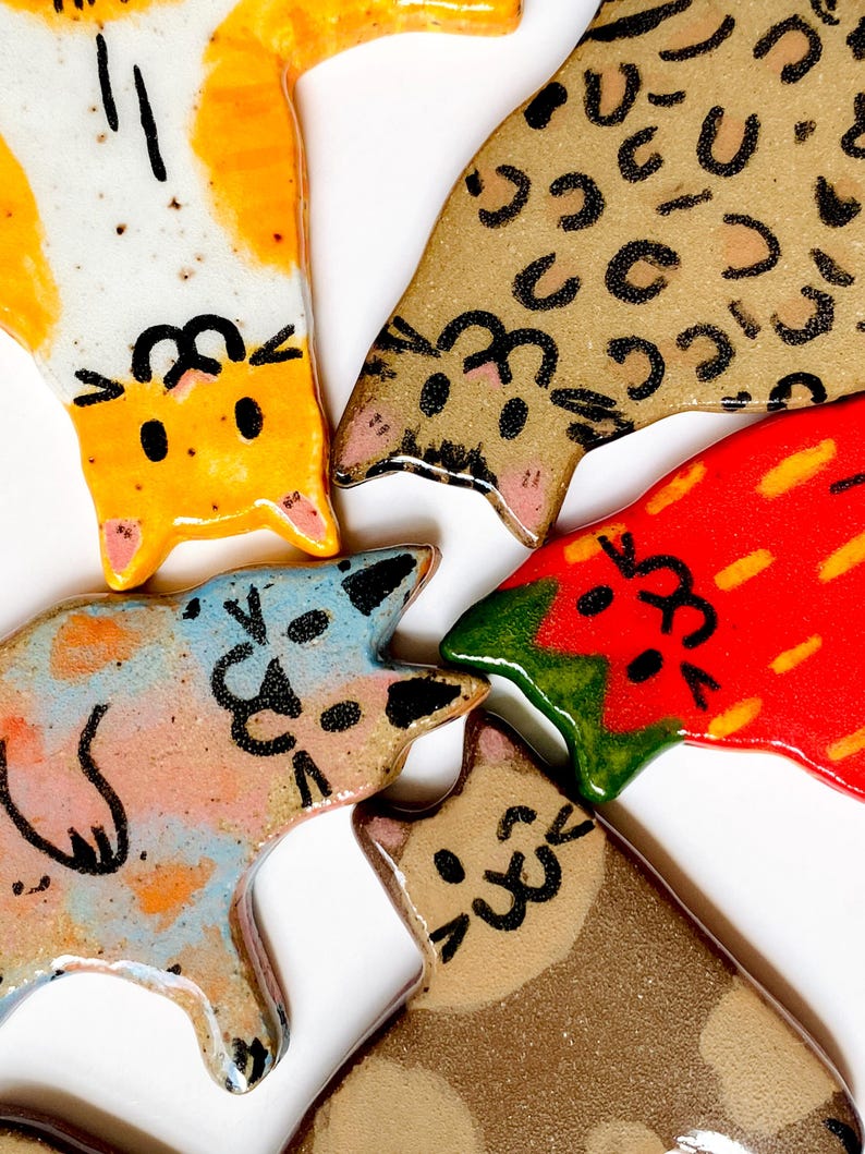 May include: Five ceramic cat-shaped ornaments with glossy finishes. One is white and orange, one is tan with black spots, one is blue and orange, one is red with yellow streaks, and one is brown with darker brown patches, all featuring black line art faces.