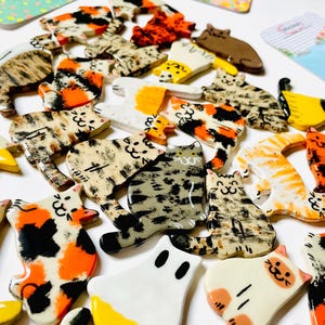 May include: A collection of small, flat, ceramic cat-shaped ornaments featuring various coat patterns including calico, tabby, solid white, and orange. Each piece displays a hand-painted feline face with simple line details and a glossy finish.