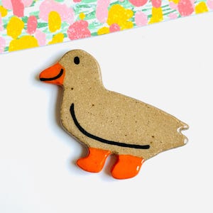 May include: A ceramic duck figurine with a speckled tan body, orange beak and feet, and a black outline. The duck is on a white surface with a colorful floral pattern in the background.