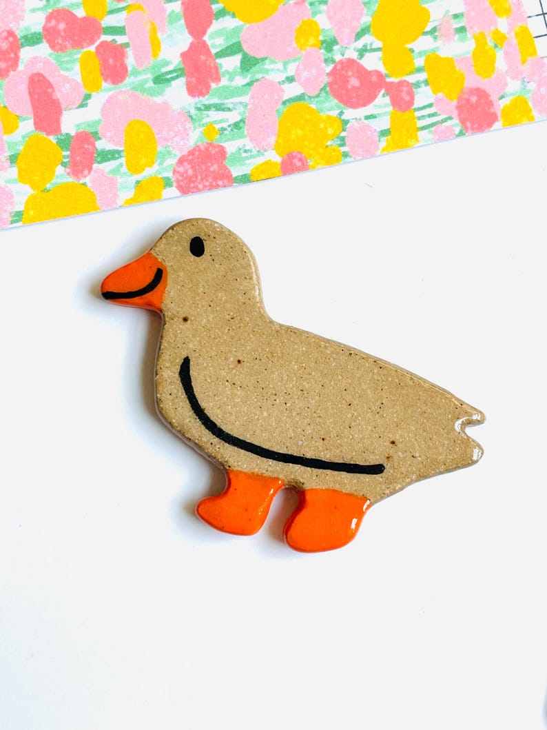 May include: A ceramic duck figurine with a speckled tan body, orange beak and feet, and a black outline. The duck is on a white surface with a colorful floral pattern in the background.
