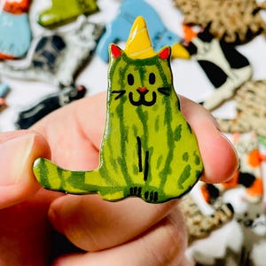 May include: A ceramic cat-shaped pin featuring a green watermelon-inspired pattern with dark green stripes. The cat has a yellow party hat, red inner ears, and a hand-painted black face with whiskers, a small red nose, and a smiling mouth.