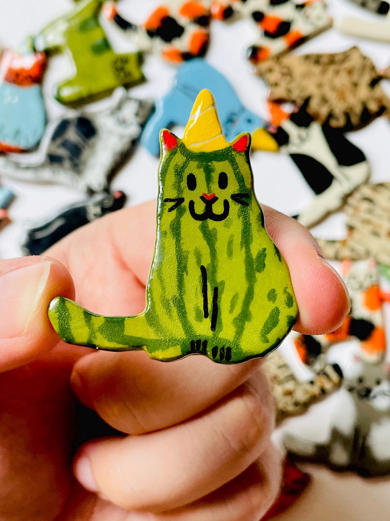 May include: A ceramic cat-shaped pin featuring a green watermelon-inspired pattern with dark green stripes. The cat has a yellow party hat, red inner ears, and a hand-painted black face with whiskers, a small red nose, and a smiling mouth.