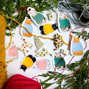 May include: A collection of colorful cat illustrations with different patterns and designs. The cats are wearing sweaters and have various expressions on their faces. The illustrations are on a white background with green foliage.