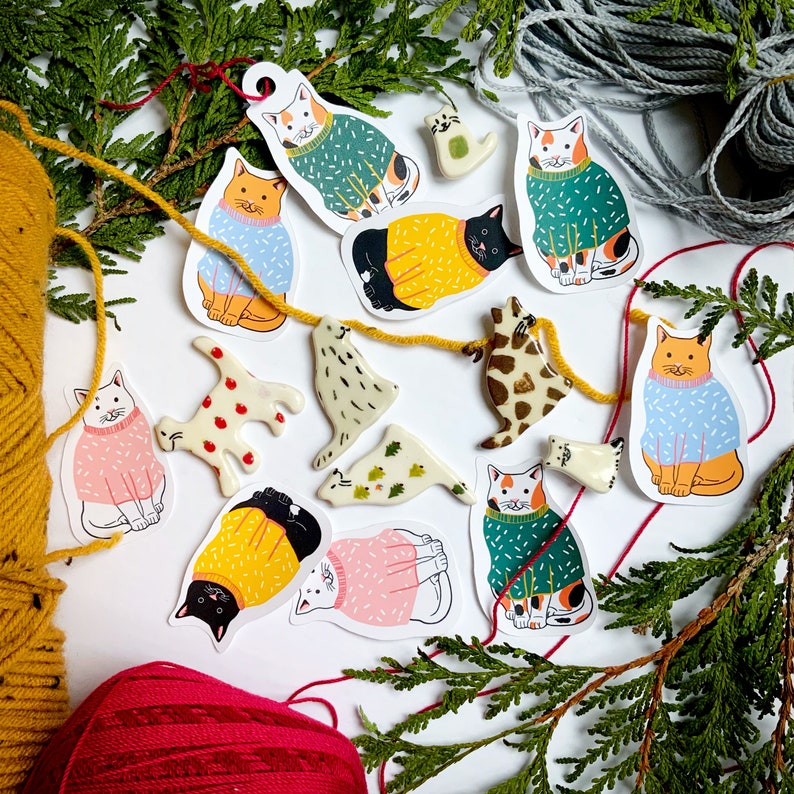 May include: A collection of colorful cat illustrations with different patterns and designs. The cats are wearing sweaters and have various expressions on their faces. The illustrations are on a white background with green foliage.